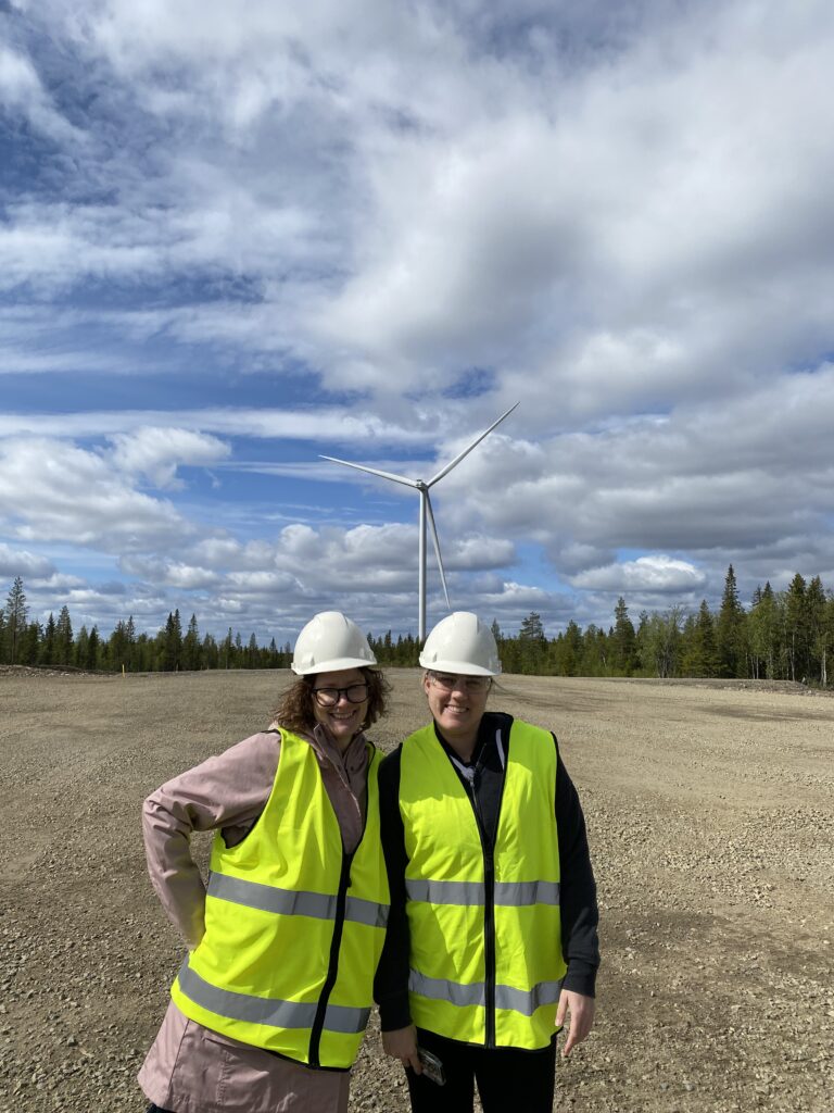 Jamie and Mallory stand in front of a giant wind mill out in the middle of Markbygden Wind Farm. 
